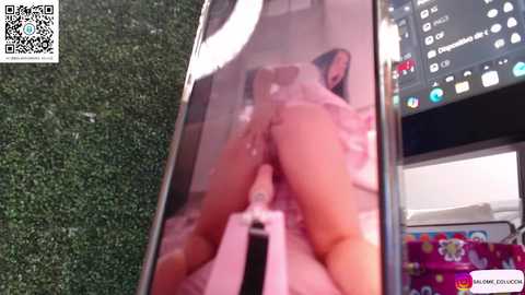 Video of a naked person's buttocks and genitals reflected in a smartphone screen, partially visible on a green grass background, with a QR code watermark.