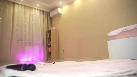 Video of a minimalist, well-lit bedroom with beige walls, white curtains, and a white bed with a white blanket. A small wooden shelf holds folded clothes, and an air conditioning unit is mounted on the wall. A black vibrator lies on the bed.