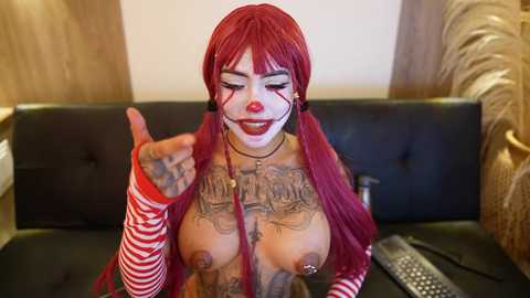 Video of a woman with red hair in a clown costume, large breasts, and multiple tattoos, standing on a black leather couch, giving a thumbs-up, indoors.