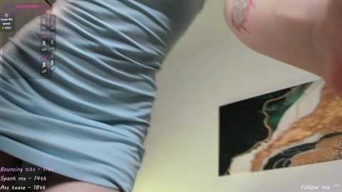 Video of a woman in a tight, blue, ruffled top, revealing her stomach and partial breasts. Her skin is pale, and she has a tattoo on her right arm. Background includes a framed picture and a TV screen showing a \"Bound It's Me\" video.