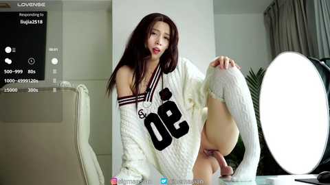 Media: Video of an Asian woman in a white, oversized sports jersey, wearing knee-high socks, seated on a table in a well-lit room with a large, round light.