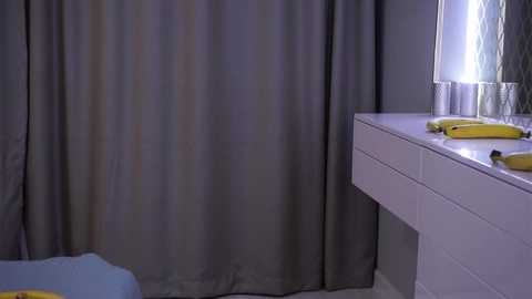 Media: A video of a minimalist bathroom featuring a white dresser with a single banana and a glass lamp on it, gray curtains, and a light blue chair in the background.