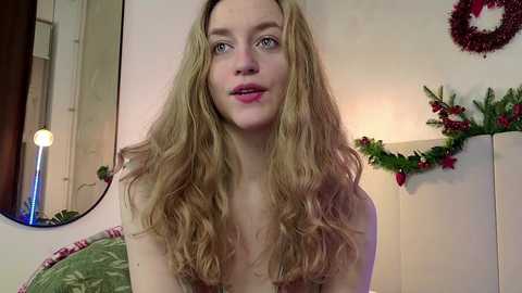 Media: Video of a young, fair-skinned woman with long, wavy blonde hair, wearing a green dress, standing in a warmly decorated room with Christmas decor, including a wreath and garland.
