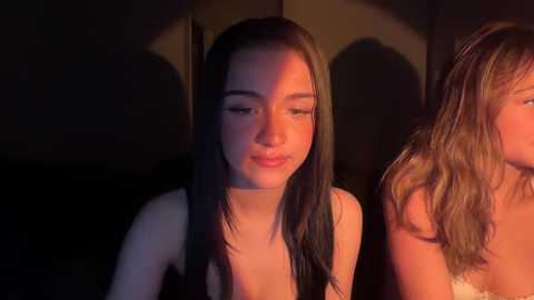 Video of two young women with fair skin, long dark hair, and light makeup, sitting close together in a dimly lit room, bathed in warm, red-hued lighting.