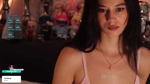 A video of a young woman with long black hair and light skin, wearing a pink spaghetti-strap top and a gold necklace. She stands in front of a dimly lit room filled with plush toys.