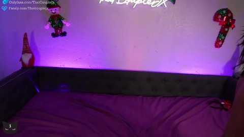 A video of a dimly-lit bedroom with a plush purple bed, illuminated by purple light. Two festive, colorful Elf on the Shelf dolls hang on the wall, and a 'Hollywood' neon sign is partially visible.