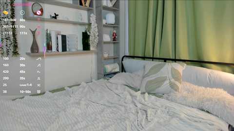 Video of a modern, minimalist bedroom with white bedding, green curtains, and a gray metal headboard. The background features a white shelf with decorative items, and the lighting is soft and ambient.