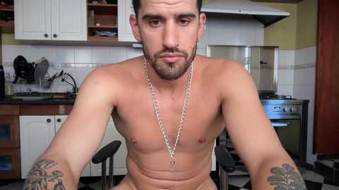 Video of a shirtless, muscular man with a beard and tattoos, wearing a silver chain, sitting in a kitchen with white cabinets and a stainless steel stove.