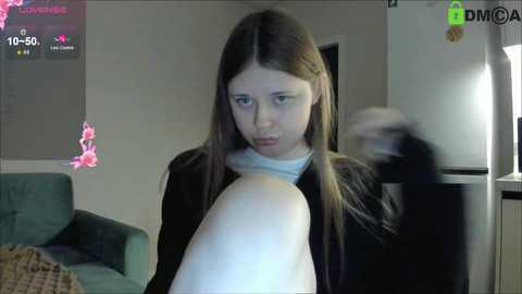 A video of a young woman with long brown hair wearing a black shirt, holding a large white pillow in a dimly lit room with a green couch, a TV, and a fridge in the background.