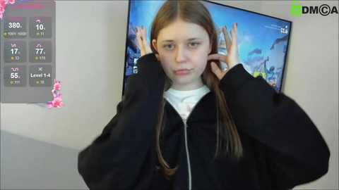 A video of a young Caucasian woman with long brown hair, wearing a black jacket, adjusting her earpiece. Background shows a TV screen displaying a weather forecast and a digital clock.