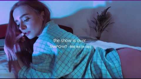 Video of a young woman with long, straight red hair, lying on a bed, covered with a blue checkered blanket, in a dimly lit room. Text overlay reads: \"the show is over. SNAPCHAT-66.6k likes.\