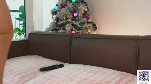 Video of a cozy living room with a brown leather sofa, a festive decorated Christmas tree with colorful lights, and a black vibrator on the light pink blanket.