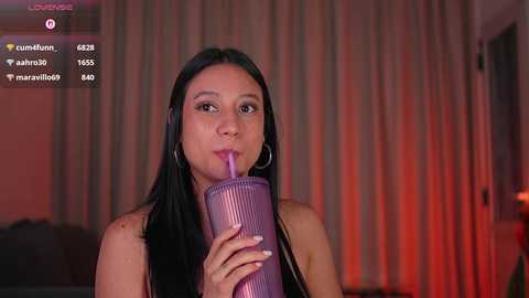 Video of a young Asian woman with long black hair, wearing hoop earrings, drinking from a purple, ribbed straw. She has a neutral expression, and the background is dimly lit with red and orange hues.