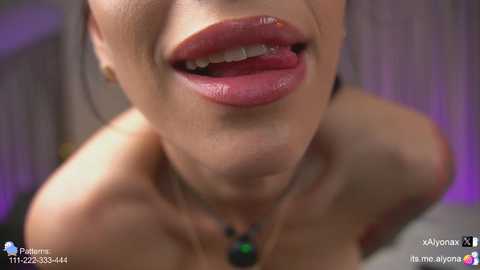 Close-up video of a woman's partially exposed, bare chest with a hint of cleavage, wearing a black necklace, and showing off a tongue piercing. Background is blurred, suggesting an indoor setting.
