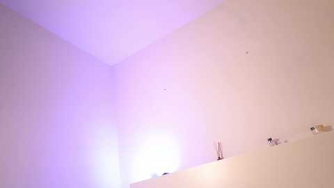 A minimalist video of a white corner room with soft purple lighting. A small table holds a few decorative items, including a candle and a small glass vase. The walls are bare, creating a clean, modern aesthetic.
