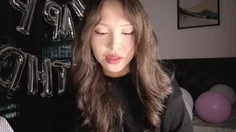 Video of an Asian woman with long, wavy brown hair, wearing a black turtleneck, blowing bubbles in a dimly lit room with black and white decor, including a framed landscape and a pink balloon.