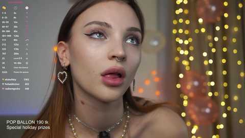 Video of a fair-skinned, blue-eyed, light-skinned woman with long, straight, auburn hair, wearing a heart-shaped nose ring, a septum ring, and a heart-shaped earring, set against a bokeh background with golden lights.