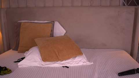 Video of a neatly made bed with two mustard-yellow and one white pillow on a beige tufted headboard. The bedding is white, and a black remote control is on the bed.