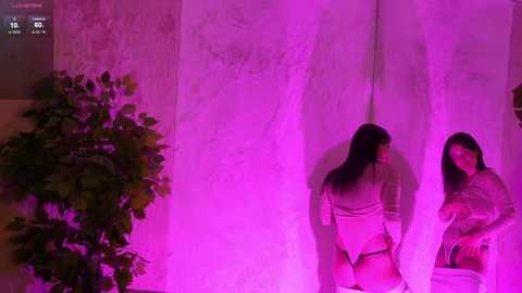 A video with a woman in a white top and black panties, standing against a textured, light-colored wall, bathed in pink light. A green plant is visible in the background.