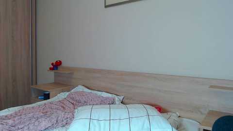 Video of a minimalist bedroom with a light wooden headboard, white pillowcases, and a pink ruffled blanket. Red toy cars are on a nightstand.