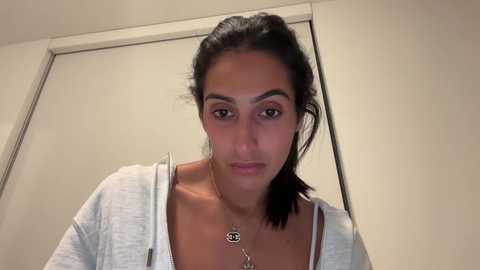 Video of a young woman with olive skin, dark hair in a ponytail, wearing a light gray hoodie, standing in front of white wardrobe doors.