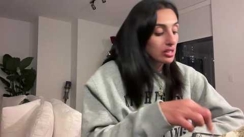 Video of a young woman with long black hair, wearing a light grey hoodie, eating in a modern, minimalistic living room.