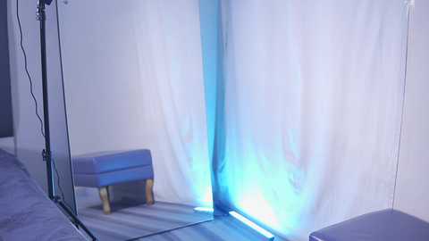 Video of a minimalist, modern room with white walls and a blue light casting a bright, vertical beam on a small, light blue bench with wooden legs. The scene is clean and industrial, with a single black power cord visible.