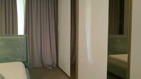 Video of a minimalist, modern bedroom with muted tones. A bed with a light green upholstered headboard is visible on the left. The walls are white, and a large window with sheer white curtains is partially covered by a light purple curtain.