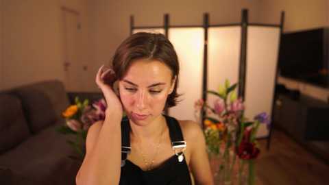 Video of a young woman with short brown hair, wearing black overalls, looking thoughtful in a warmly lit living room with a floral arrangement and a grey sofa in the background.