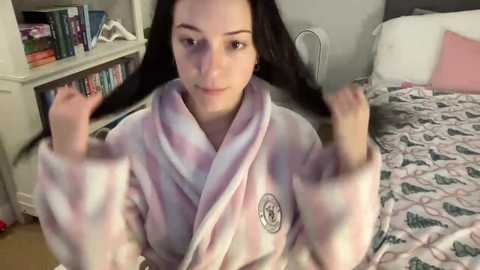 A video of a young woman with long black hair, wearing a pink and white plaid robe, sitting on a bed with a patterned duvet, in a cluttered bedroom.