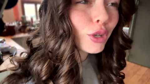 Video of a fair-skinned woman with voluminous, wavy brown hair, wearing a light grey top, in a modern kitchen with wooden floors and cabinets.
