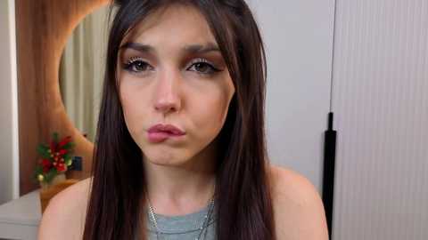 Video of a young woman with fair skin, straight brown hair, wearing a grey top and a silver necklace, looking slightly pouty, with a round mirror and a floral arrangement in the background.