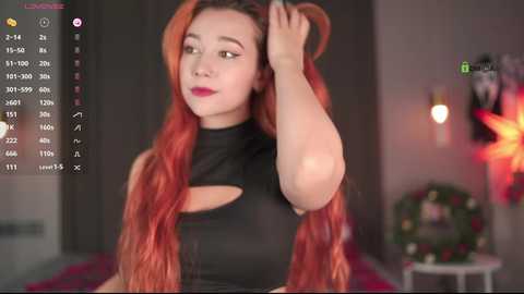 Video of a young woman with long, vibrant red hair, wearing a black, sleeveless crop top, in a dimly lit room with a festive wreath and Christmas decorations in the background.