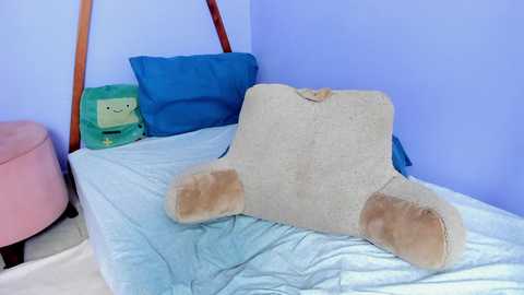 A video of a small, beige plush toy resembling a person, lying on a bed with a blue and white quilt. The room has light blue walls and a pink ottoman in the background.