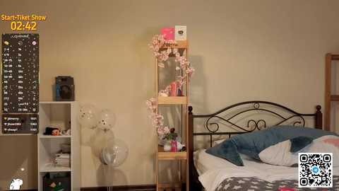 A video of a cozy, dimly lit bedroom with a white bed, teal bedding, and a wooden bookshelf adorned with pink flowers.