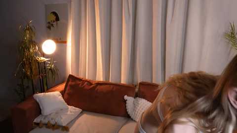 Video of a cozy living room with beige curtains, orange sofa, white pillows, a round lamp, and a person's long hair in the foreground.