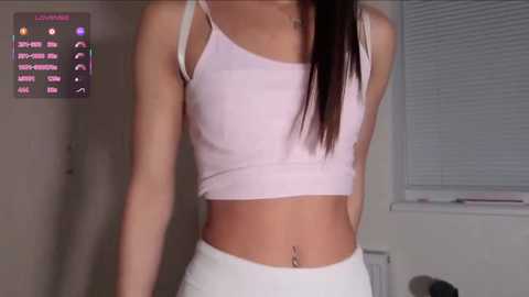 Video of a slender, light-skinned woman with long dark hair, wearing a white tank top and white pants, standing indoors against a window with blinds.