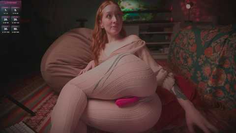 Video of a fair-skinned, red-haired woman in a beige ribbed sweater and matching pants, lounging on a beanbag chair in a dimly lit room with colorful, patterned pillows and a TV in the background.