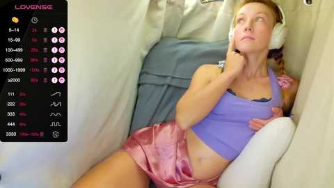 A video of a blonde woman in a purple crop top and pink shorts, wearing headphones, reclining in a plane seat, looking thoughtful.