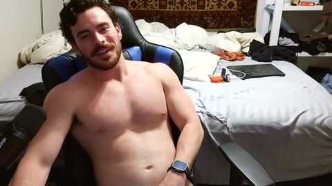 Video of a shirtless, muscular, white man with curly brown hair and a beard, sitting on a blue gaming chair in a messy bedroom.