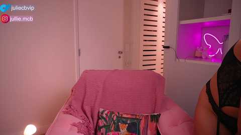 Video of a woman in black lace lingerie, lounging on a pink bed with floral pillow, in a dimly lit, pink-lit bedroom with a neon sign.