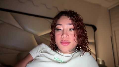 Video of a young woman with curly, shoulder-length brown hair and light skin, wearing a light gray t-shirt. She stands indoors with a bed and wardrobe in the background.