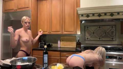 Video of two topless women in a modern kitchen; one blonde woman with medium breasts holds a wine glass, the other with long blonde hair bends over a counter.