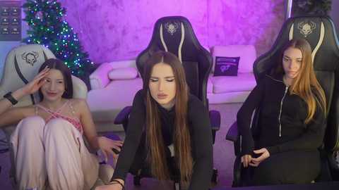 Video of three young women in a cozy living room, with one seated in a black gaming chair, wearing a black hoodie, and the other two in casual attire, seated on a couch, under purple lighting.