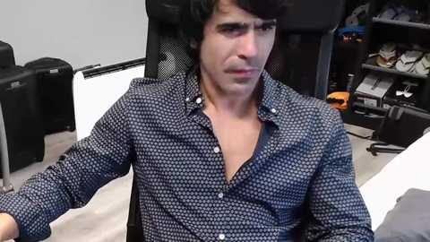Video of a man with short dark hair, wearing a blue polka-dot button-up shirt, sitting in a black ergonomic office chair in a cluttered, dimly lit room.
