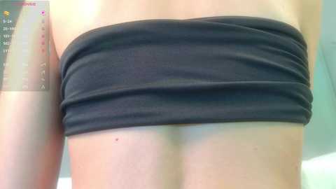 Video of a woman's bare back with a strapless, black bandeau top. The background shows a blurry, light-colored room with a partially visible screen displaying a weather forecast.