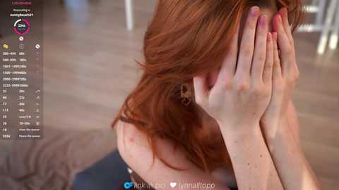 Video of a fair-skinned woman with long, reddish-brown hair, covering her face with her hands, sitting on a couch in a living room. Background shows a blurred wooden floor and furniture.