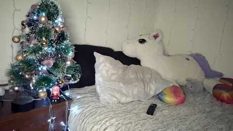 Video of a cozy bedroom with a small Christmas tree adorned with baubles, fairy lights, and a plush unicorn lying on a bed, surrounded by white bedding and a colorful pillow.