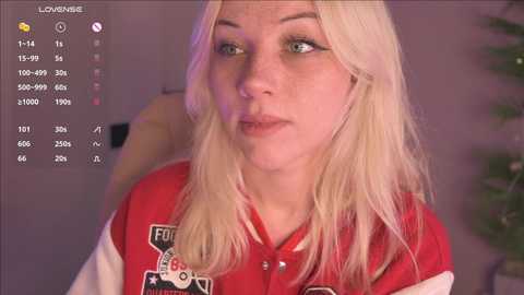 A video of a young, fair-skinned woman with long, straight blonde hair, wearing a red and white varsity jacket, looking contemplative. The background is blurred with a potted plant and a wall.