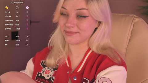 A video of a young blonde woman with a light complexion, wearing a red and white varsity jacket, seated in a beige leather chair. The background shows a dimly lit room with a door and a video recording interface.